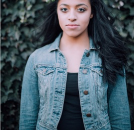 Amie S stands in front of a vertical surface covered in leaves. Amie has a medium skintone with black hair, and wears an unbuttoned light blue denim jacket with a black top underneath.