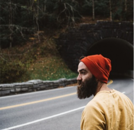 Andy C stands in front of a road that curves left into a tunnel in a hill. Andy has light skintone and a trimmed, bushy brown beard. Andy faces the viewer's left, wearing a burnt red beanie and mustard yellow long-sleeve shirt.