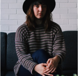 Angela W sits cross-legged on a dark grey couch in front of a white brick wall, hands resting on their right leg. Angela has shoulder length chestnut hair, pale skin and wears a striped grey-brown jumper.