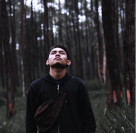 Brian M stands in a sparse forest, head tilted away from the camera towards the sky. Brian appears East Asian, has brown-black hair, and wears a black zip up hoodie with a black bum bag.