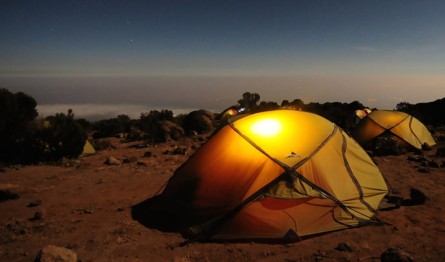 Campsite on a mountain at dusk. There are endless clouds out to the horizon. The tents shine from the lanterns turned on the inside.